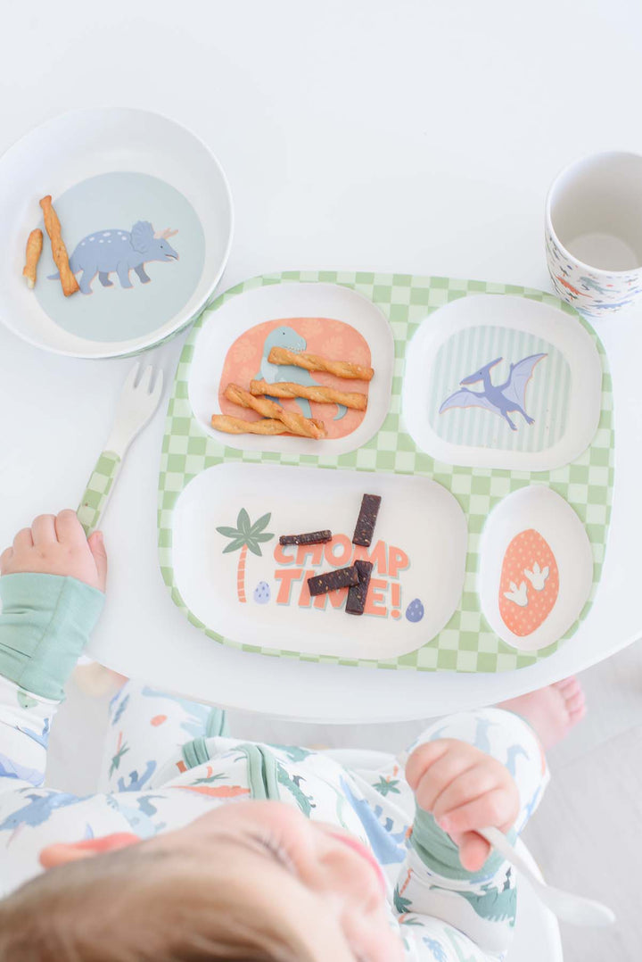 A baby in a patterned onesie sits at a white high chair table, looking down at a green and white checkered divided meal tray with dinosaur and palm tree illustrations. The tray contains snacks, next to a matching bowl and a white plastic fork.