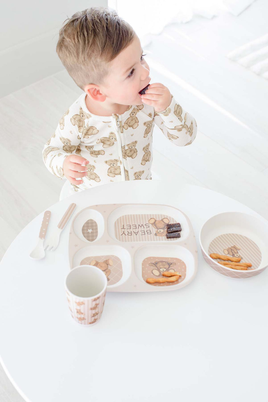 A toddler wearing a Caden Lane cream long-sleeve pajama with a brown teddy bear pattern sits at a white table, eating from a Caden Lane matching beige compartment tray with a small cup and bowl.