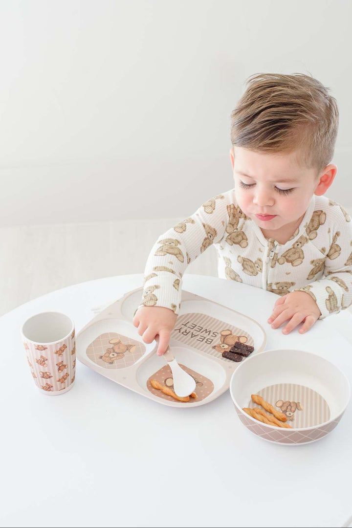 A young child wearing Caden Lane cream pajamas with a brown animal print sits at a white table, reaching for food on a divided tray. A matching Caden Lane patterned cup and bowl are also on the table.