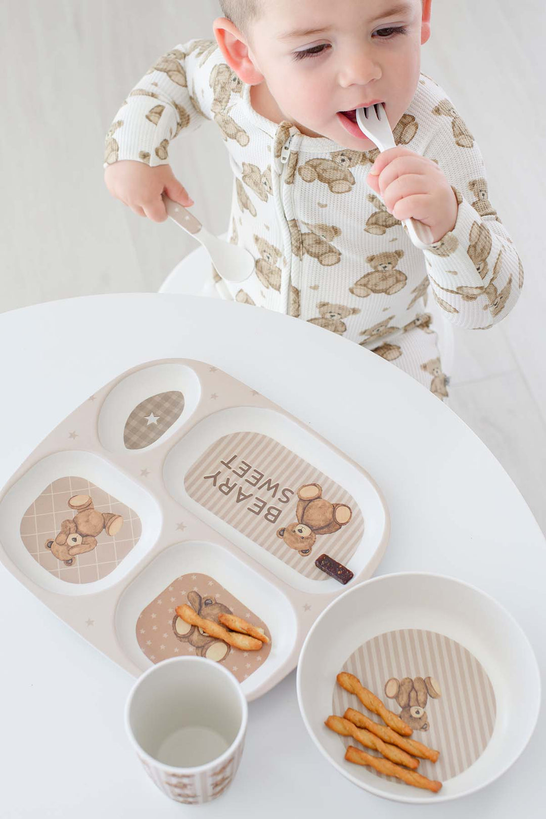 A baby wearing a Caden Lane white and beige teddy bear print footed sleeper sits at a white table, using a Caden Lane matching beige divided tray with snacks and a small cup.