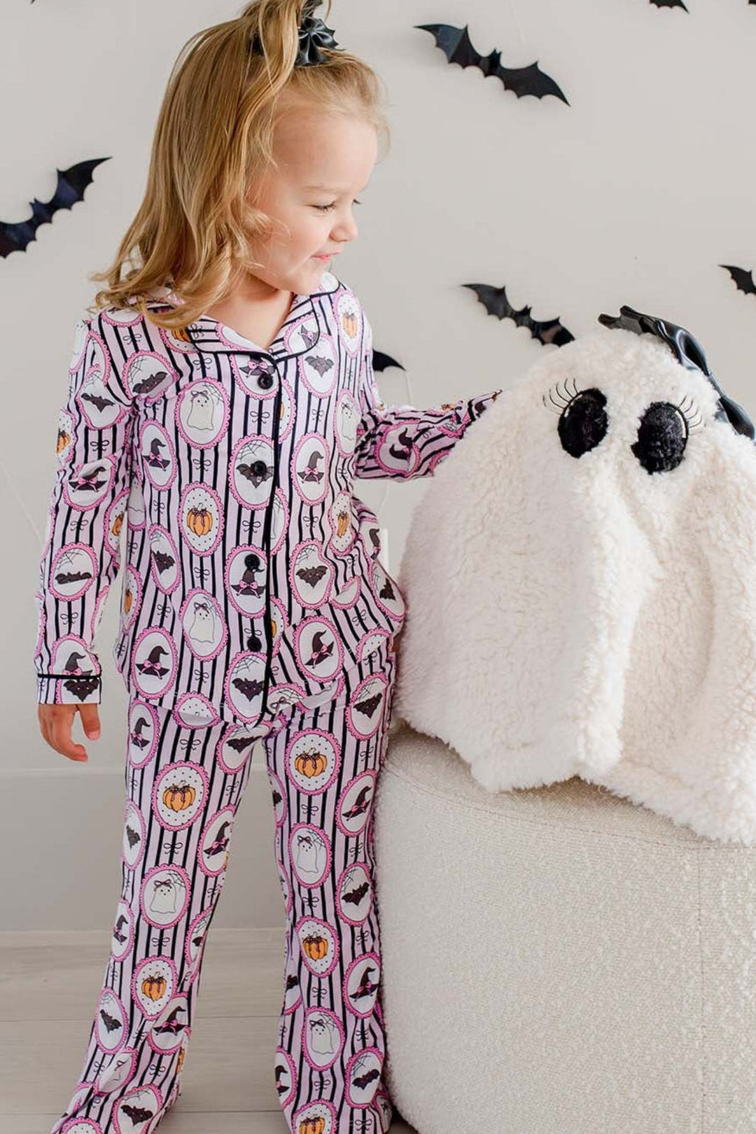 Girl standing next to plush ghost and wearing Caden Lane pink and black striped Halloween pajamas with ghost, bat, and pumpkin cameo print button-up two-piece set.