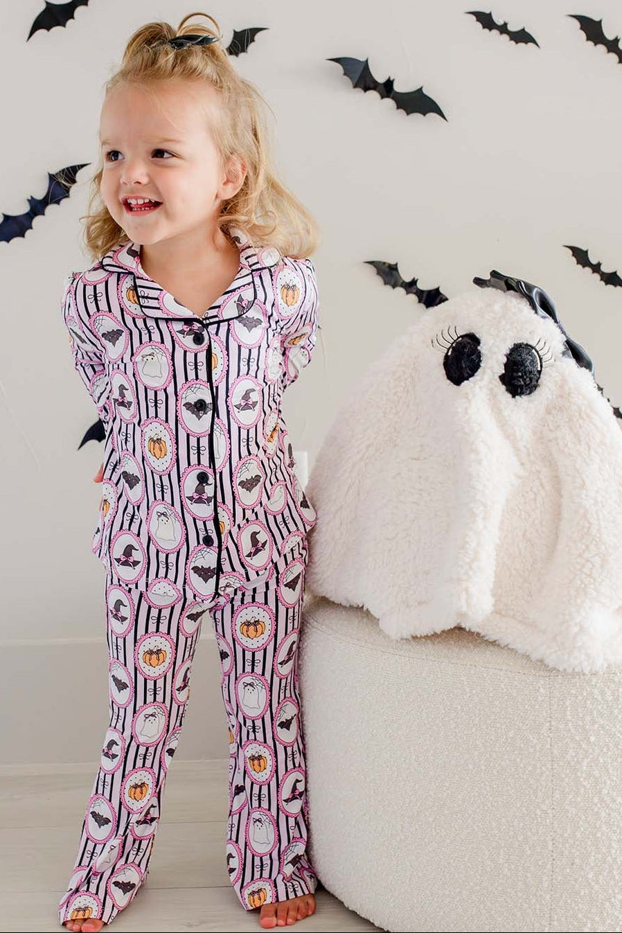 Young girl standing next to a plush ghost-shaped ottoman cover, wearing a Caden Lane pink and black striped button-up two-piece flare pajama set with ghost, bat, and pumpkin cameo print, with bat decorations on the wall behind her.