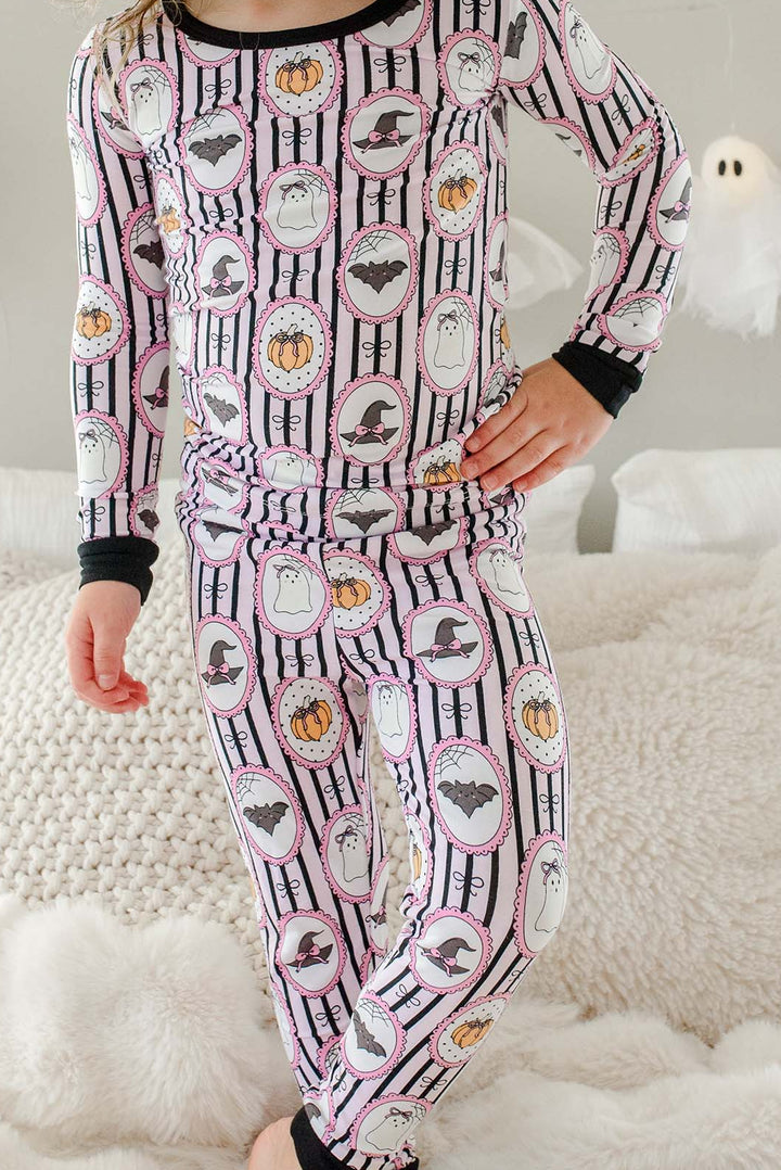 Young girl standing on a bed wearing a Caden Lane pink and black striped two-piece pajama set with ghost, bat, and pumpkin cameo print, with a black bow in her hair and hanging ghost lights in the background.