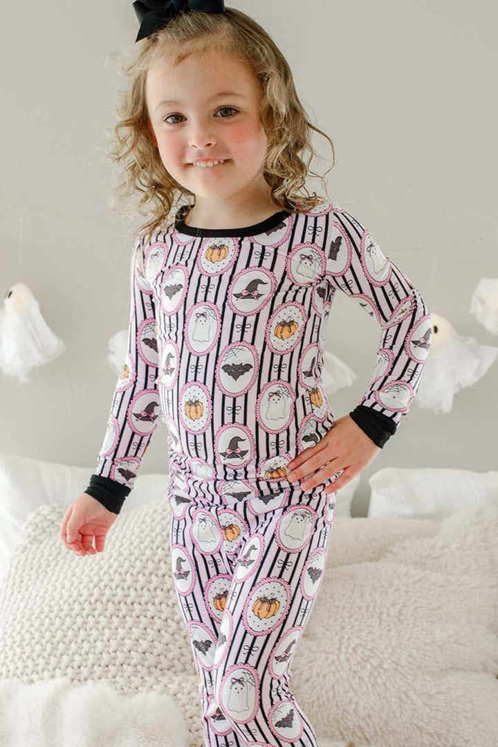 Young girl standing on a bed wearing a Caden Lane pink and black striped two-piece pajama set with ghost, bat, and pumpkin cameo print, with a black bow in her hair and hanging ghost lights in the background.
