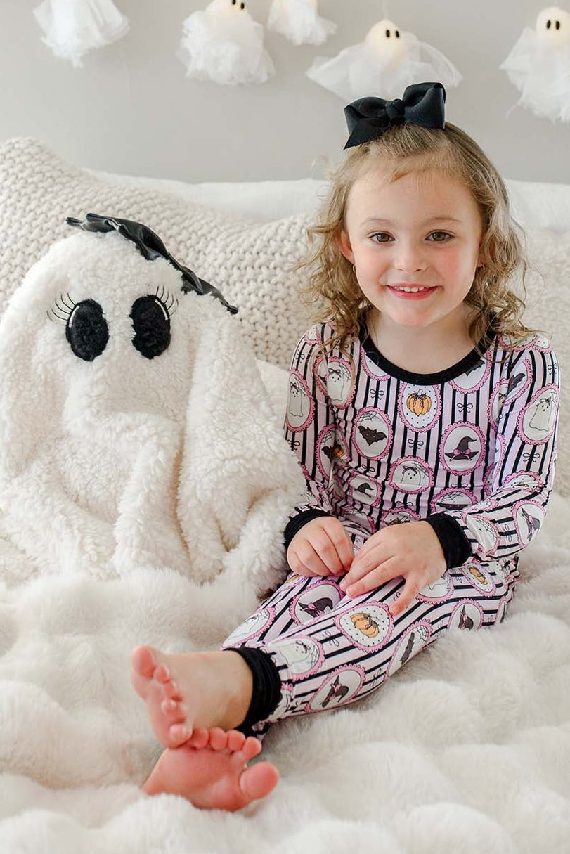 A young girl with a black bow sits on a fluffy white bed wearing a purple and white patterned Halloween-themed pajama set. A large white plush ghost toy is next to her, with a white wall and ghost string lights in the background.