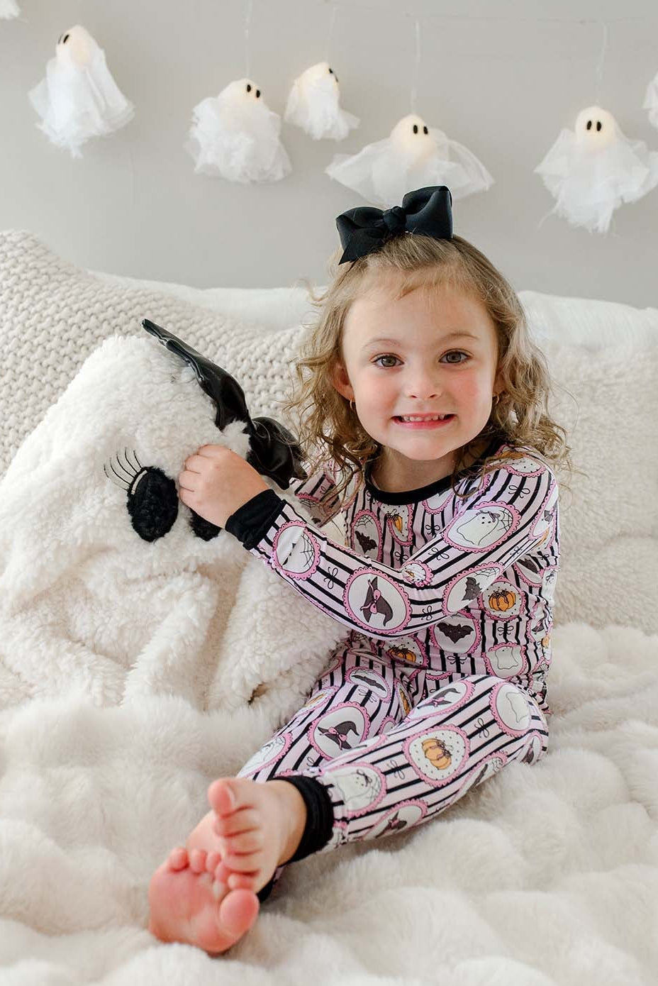 Young girl sitting on a bed wearing a Caden Lane pink and black striped two-piece pajama set with ghost, bat, and pumpkin cameo print, with a black bow in her hair and hanging ghost lights in the background.