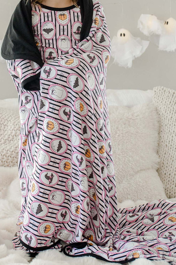 Child wrapped in a Caden Lane pink and black striped bamboo blanket with ghost, bat, pumpkin, and witch hat cameo print, standing on a couch with hanging ghost lights in the background