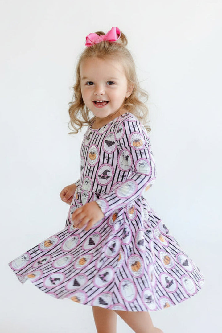 A young girl with blonde hair and a pink bow stands barefoot in a bright bedroom, wearing a long-sleeve purple twirl dress with a white striped Halloween pattern featuring pumpkins, bats, and witch hats.