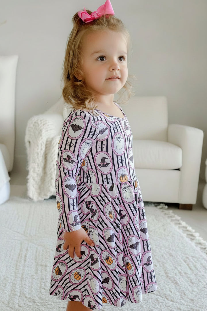 A young girl with blonde hair and a pink bow stands in a bright bedroom, wearing a long-sleeve purple twirl dress with a white striped Halloween pattern featuring pumpkins, bats, and witch hats.