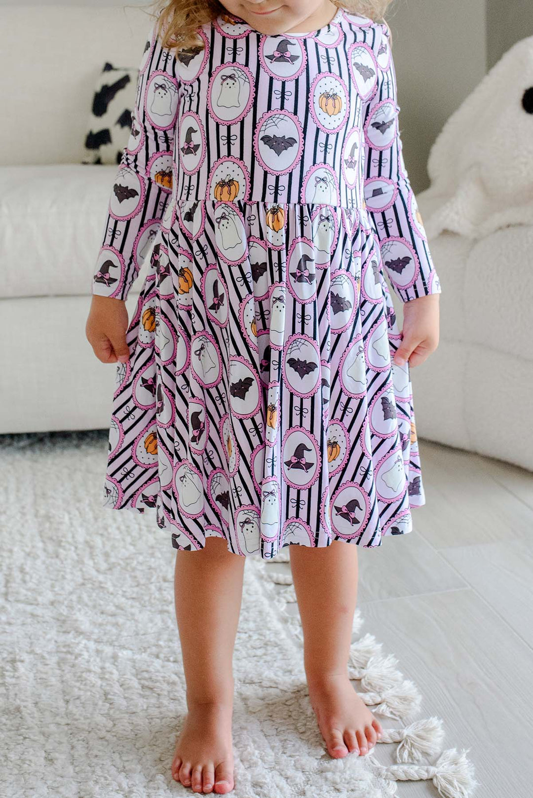 A young girl with blonde hair and a pink bow stands barefoot in a bright bedroom, wearing a long-sleeve purple twirl dress with a white striped Halloween pattern featuring pumpkins, bats, and witch hats.