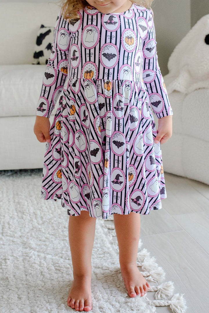 A young girl with blonde hair and a pink bow stands barefoot in a bright bedroom, wearing a long-sleeve purple twirl dress with a white striped Halloween pattern featuring pumpkins, bats, and witch hats.