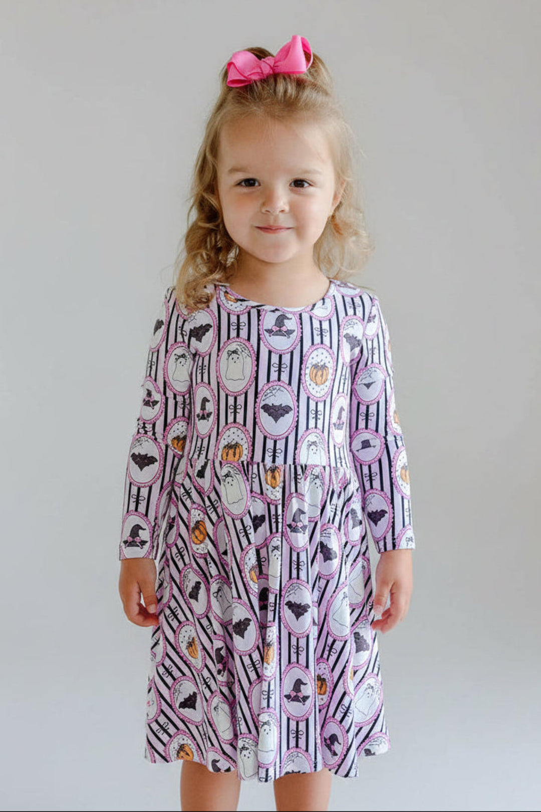 A toddler girl stands barefoot on a white rug in a bright living room, wearing a long-sleeve dress with a purple, white, and black witch-themed pattern of cauldrons, brooms, and hats. She has a pink bow in her hair.