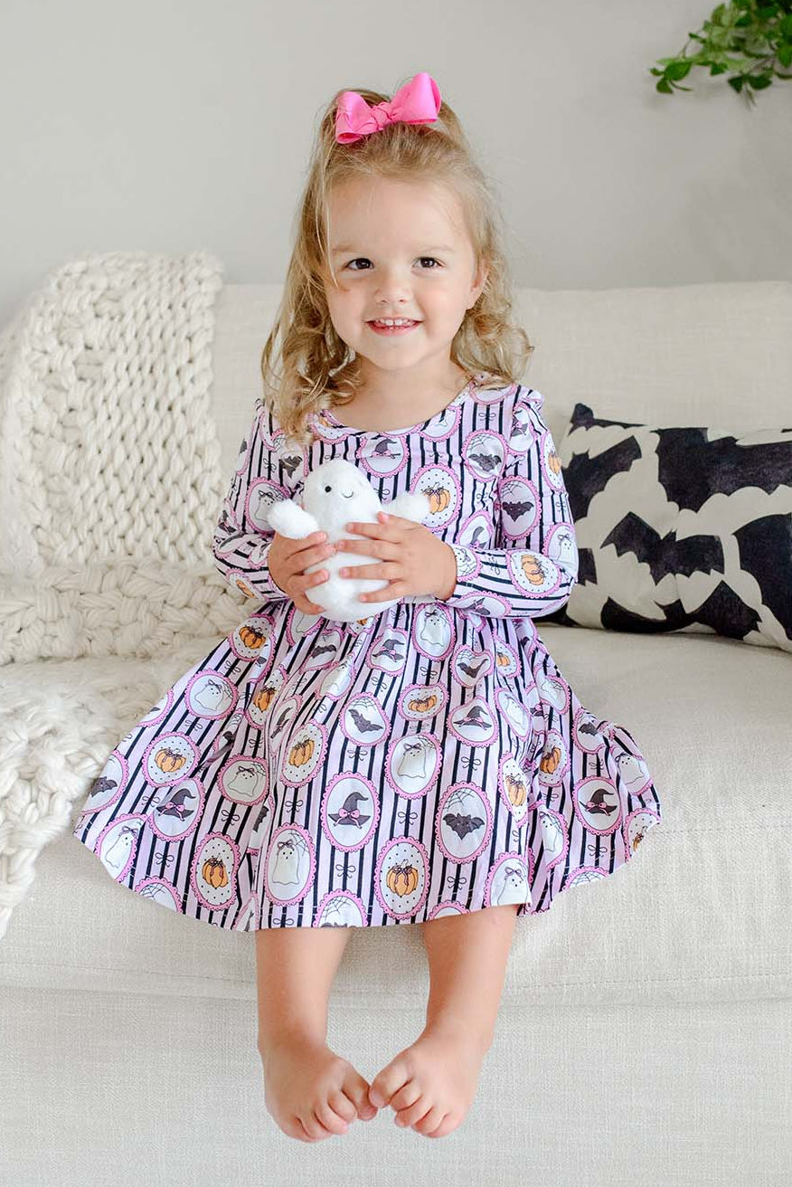 A young girl sitting on a couch holding a plush ghost toy, wearing a purple and white striped Halloween-print twirl dress with pumpkin, bat, and witch hat designs.