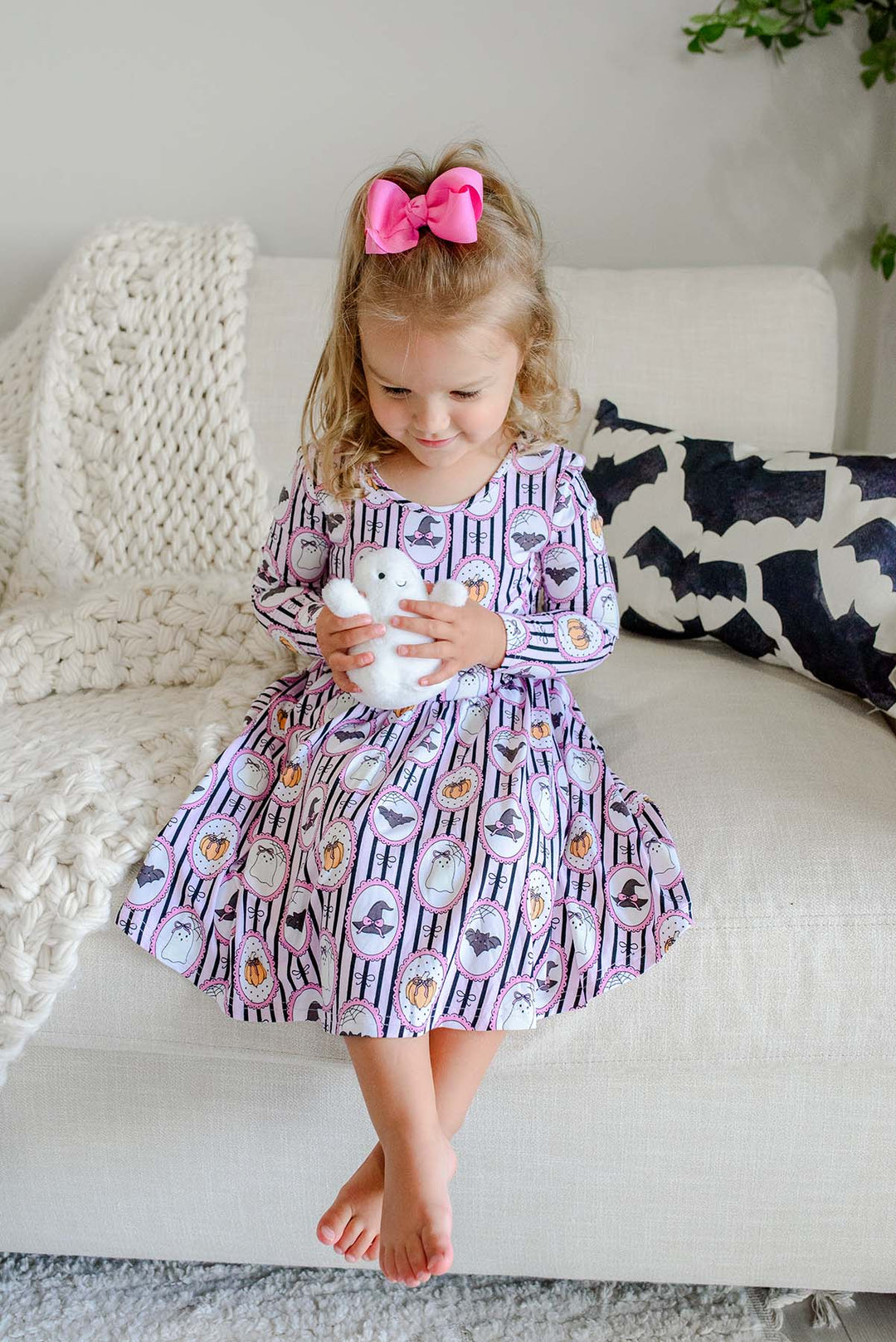 A young girl holding a stuffed ghost and wearing a pink bow sits barefoot in a bright bedroom, wearing a long-sleeve purple twirl dress with a white striped Halloween pattern featuring pumpkins, bats, and witch hats.