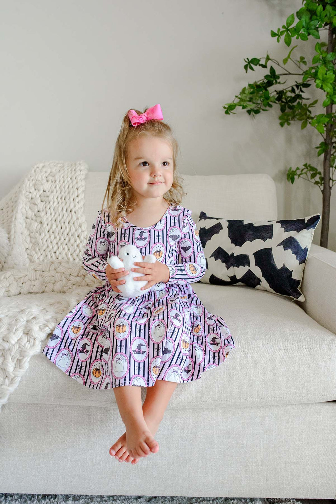 A young girl holding a stuffed ghost and wearing a pink bow sits barefoot in a bright bedroom, wearing a long-sleeve purple twirl dress with a white striped Halloween pattern featuring pumpkins, bats, and witch hats.