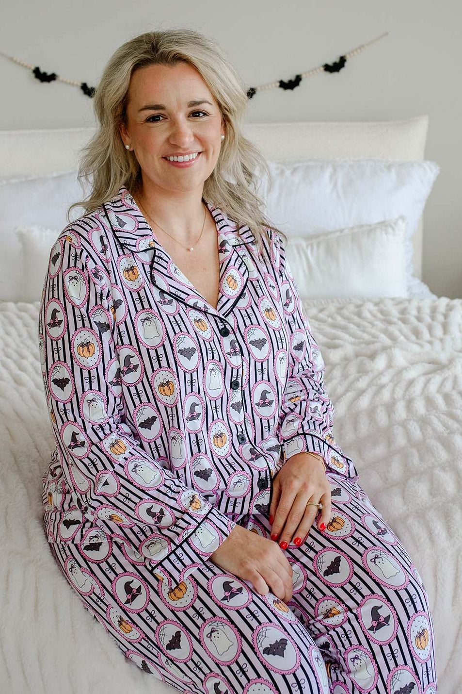 A woman wearing a matching long-sleeve pajama set with a Halloween-themed pattern, sitting on a white bed with white pillows and a white textured quilt.
