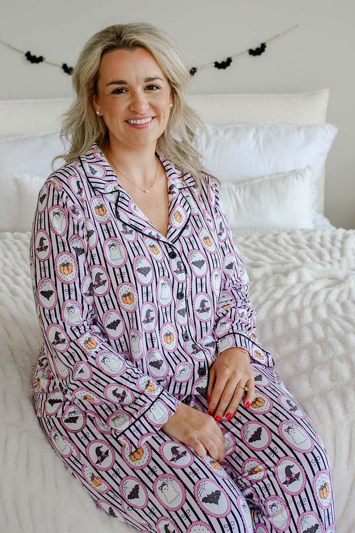 A woman wearing a matching long-sleeve pajama set with a Halloween-themed pattern, sitting on a white bed with white pillows and a white textured quilt.