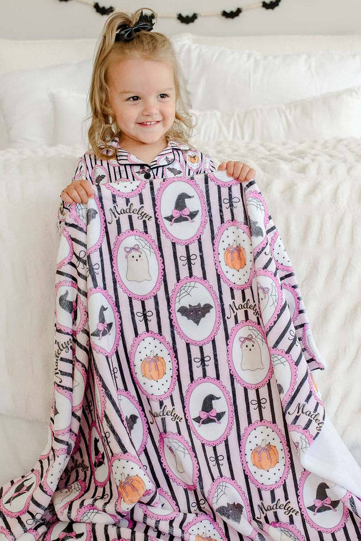 A young girl sitting on a white bed, holding up a pink and black patterned blanket with Halloween-themed illustrations like ghosts, pumpkins, bats, and witch hats.