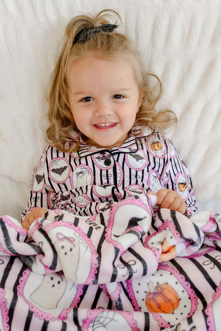 A young girl lying on a white bed, smiling and wrapped in a pink and black Halloween blanket with ghost, bat, and pumpkin designs, wearing a matching pajama top.