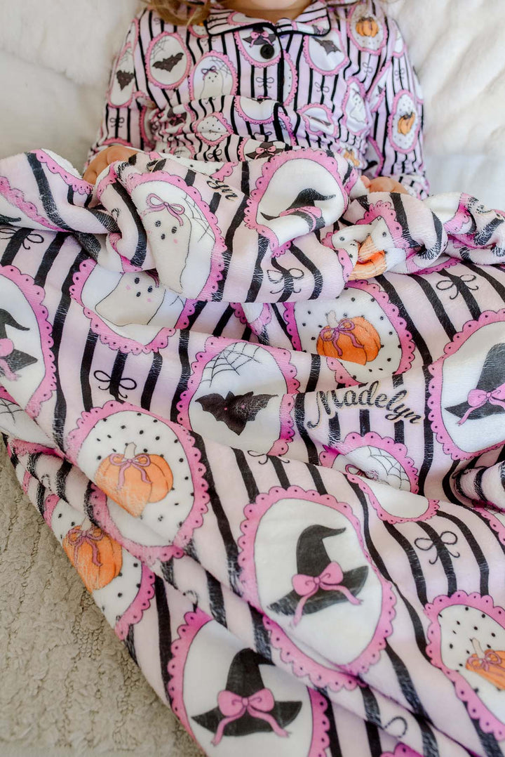 A close-up of a baby blanket and matching long-sleeve shirt featuring a Halloween print with black and white stripes, pink-trimmed ghost and witch motifs, orange pumpkins, and the name 'Madelyn'.