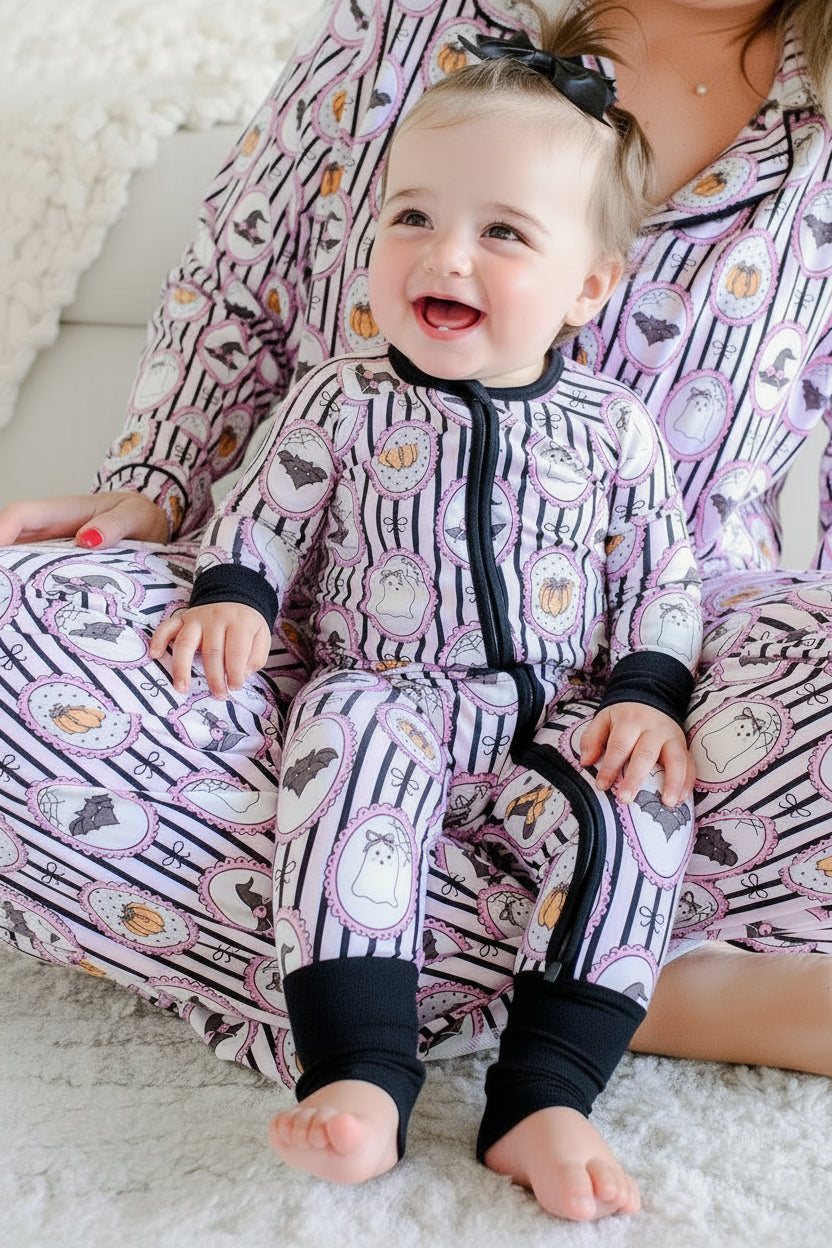 Baby sitting in an adult's lap, both wearing matching Caden Lane pink and black striped Halloween pajamas with ghost, bat, and pumpkin cameo print, the baby's romper with black cuffs and a black bow.