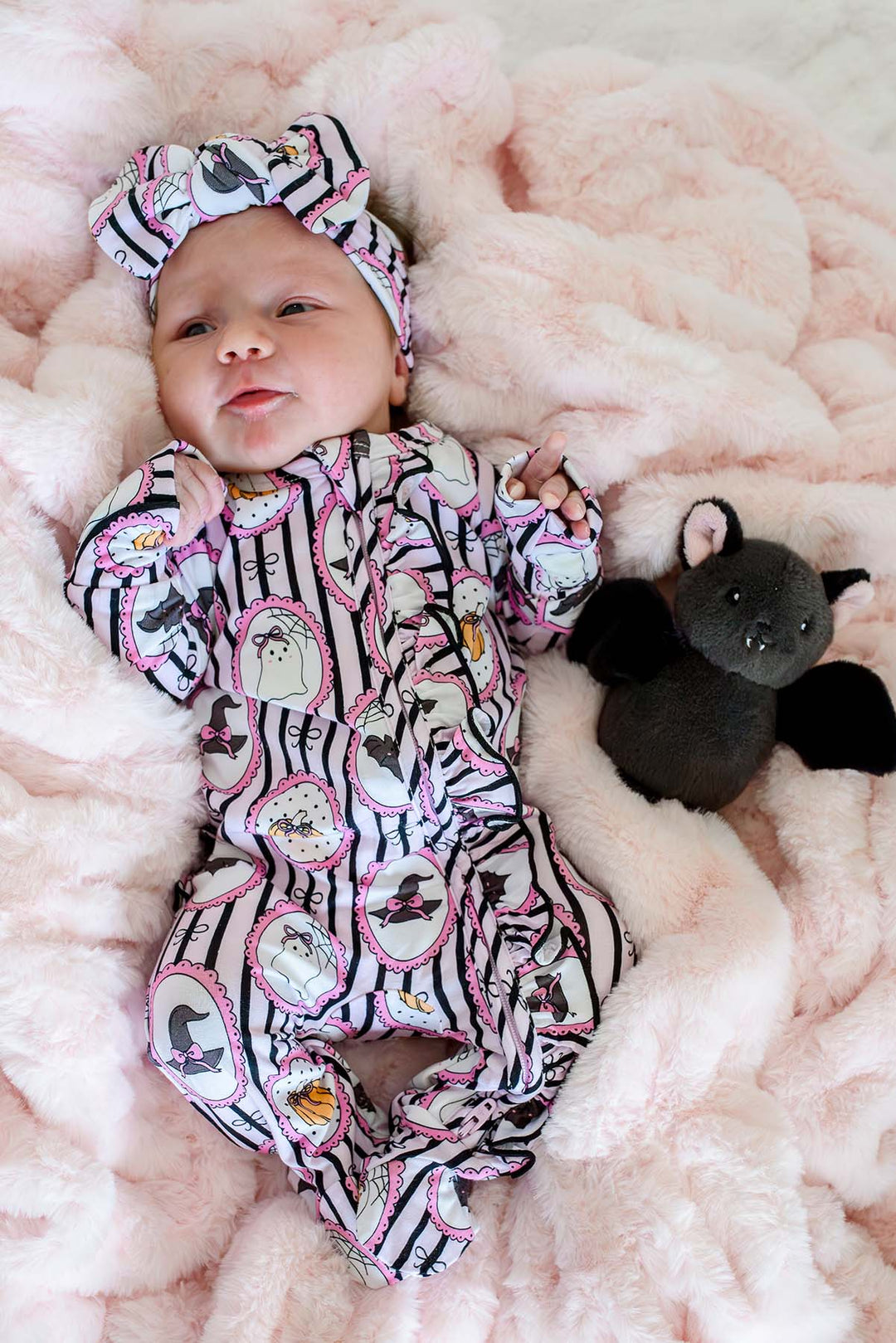 A baby wearing a pink and black witch-themed footed onesie with a matching headband, lying on a fluffy pink blanket next to a small black plush bat toy.
