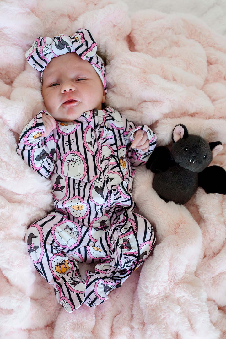 Sleeping newborn wearing a Caden Lane pink and black striped Halloween ruffle footie with ghost, pumpkin, and witch hat frame print, plus a matching bow headband, lying next to a plush black bat toy