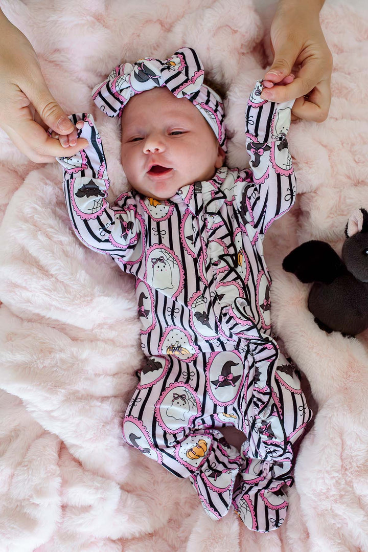 Newborn wearing a Caden Lane pink and black striped Halloween ruffle footie with ghost, pumpkin, and witch hat frame print, plus a matching bow headband, lying next to a plush black bat toy