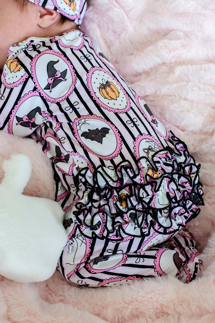 A baby sleeping on a pink plush blanket, wearing a matching Halloween-themed bodysuit and headband. The outfit features black and white stripes with pink circular borders, black bats, pumpkins, and ghosts.