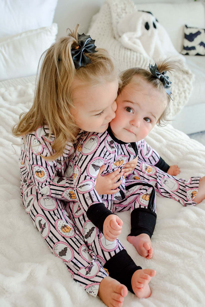 Two sisters lying together on a bed wearing matching Caden Lane pink and black striped Halloween pajamas with ghost, bat, and pumpkin cameo print — the younger in a zip-up footie and the older in a button-up two-piece set.
