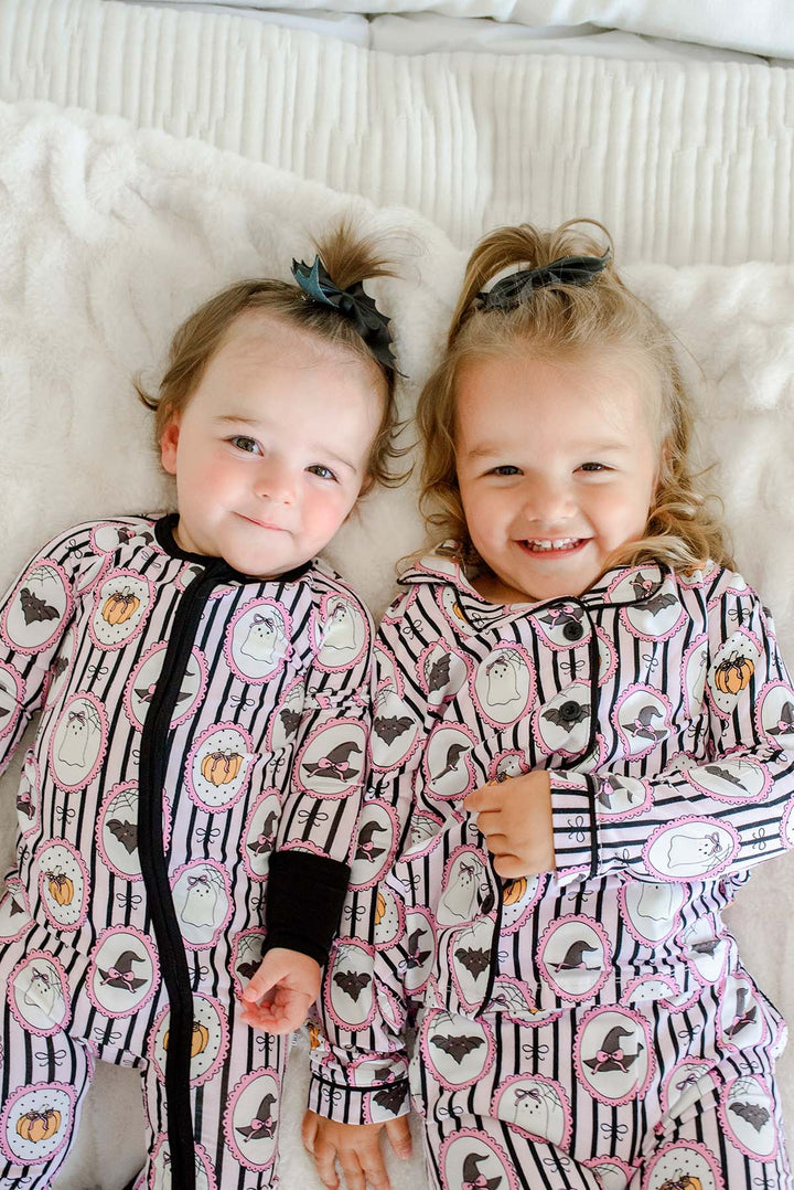 Two sisters lying together on a bed wearing matching Caden Lane pink and black striped Halloween pajamas with ghost, bat, and pumpkin cameo print — the younger in a zip-up footie and the older in a button-up two-piece set.