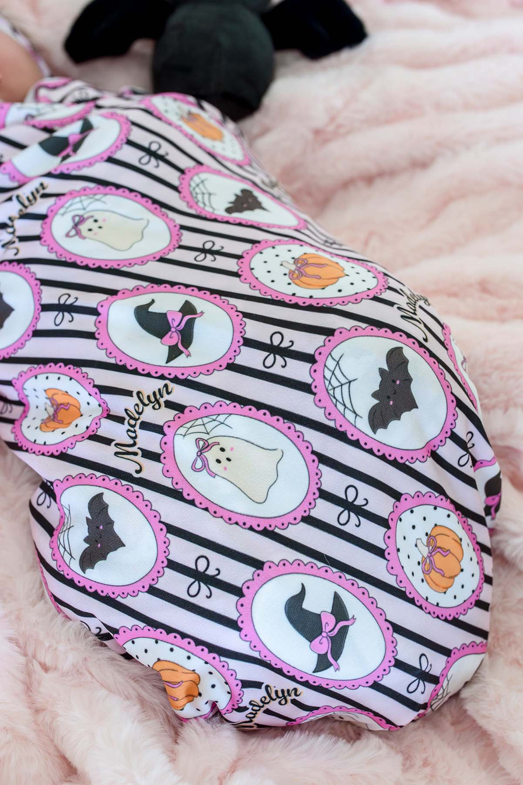 Close-up of a baby swaddled in a personalized pink and black Halloween blanket with witch hat, bat, ghost, and pumpkin designs and the name 'Madelyn'.