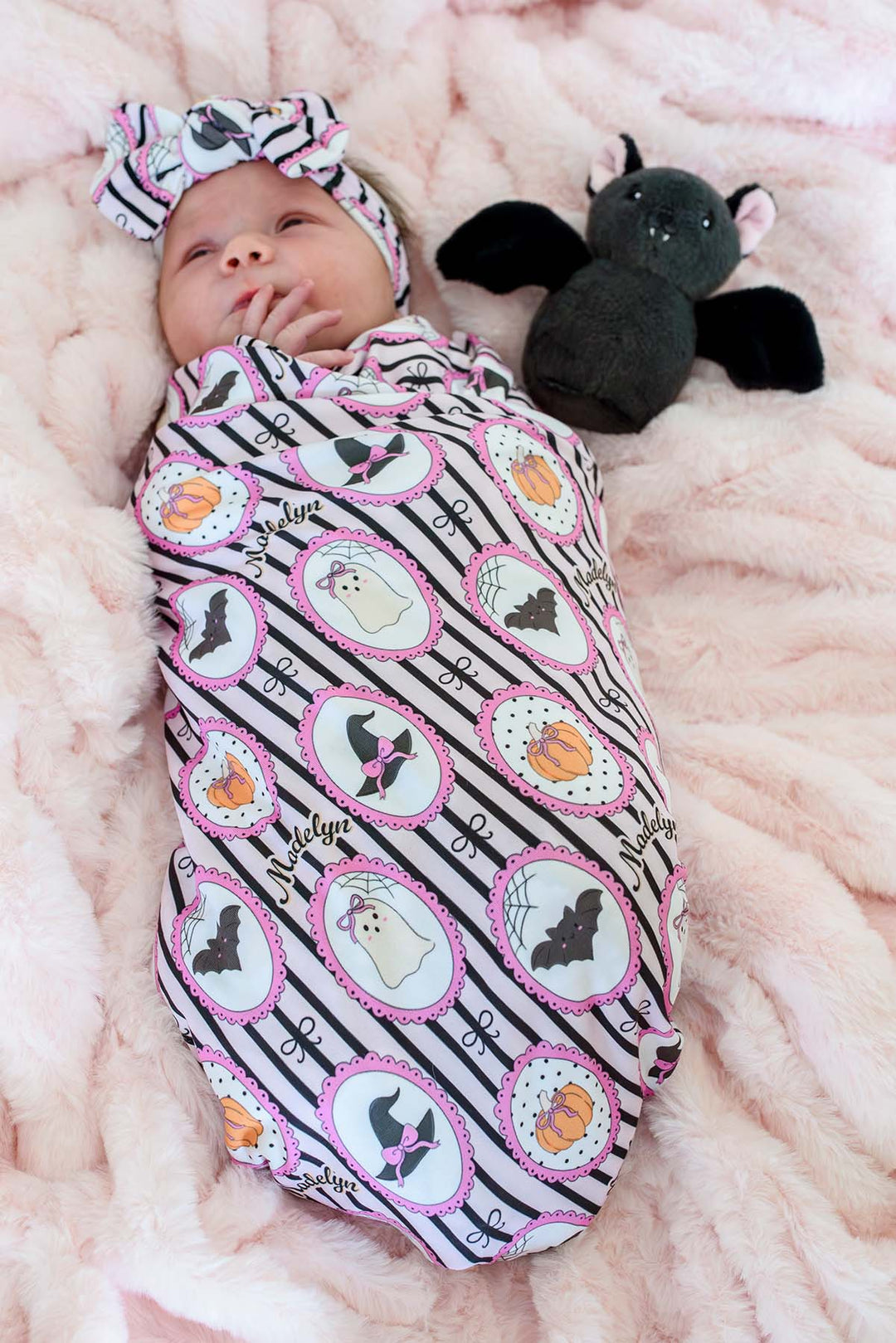 A baby swaddled in a personalized pink and black Halloween blanket with matching headband, lying on a pink blanket next to a plush bat toy.