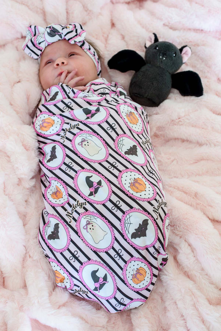 A baby swaddled in a personalized pink and black Halloween blanket with matching headband, lying on a pink blanket next to a plush bat toy.