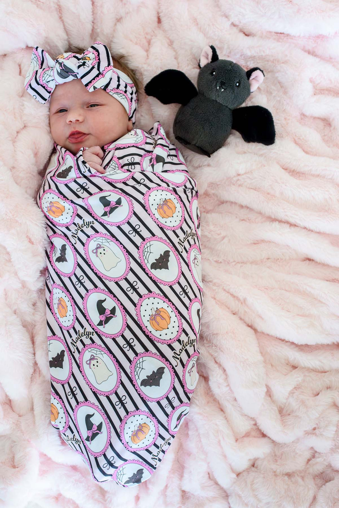 A baby swaddled in a personalized pink and black Halloween blanket with matching headband, lying on a pink blanket next to a plush bat toy.