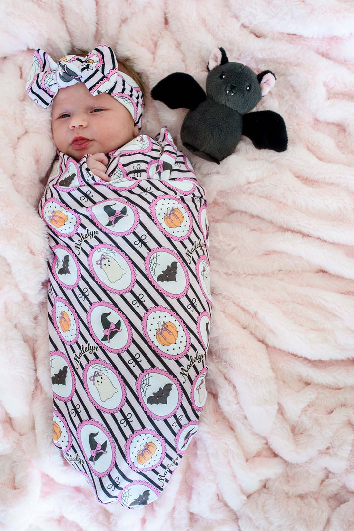 A baby swaddled in a personalized pink and black Halloween blanket with matching headband, lying on a pink blanket next to a plush bat toy.