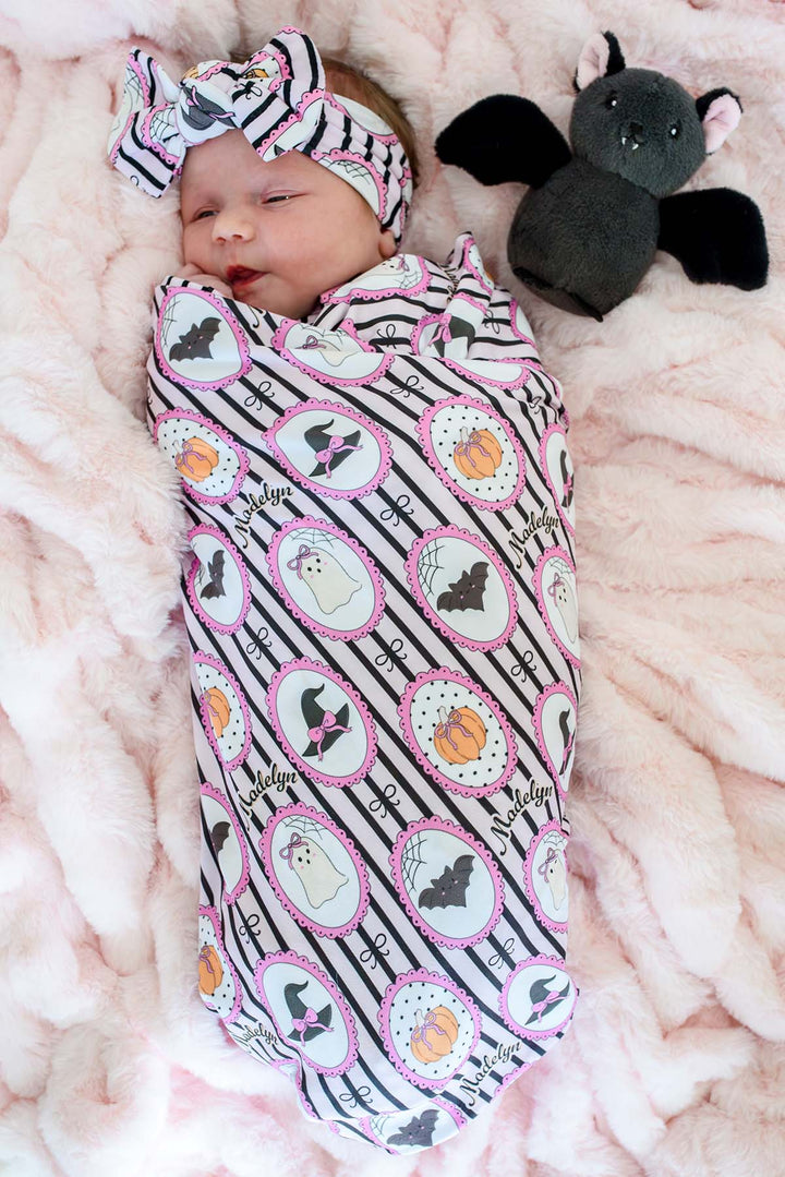 A baby swaddled in a personalized pink and black Halloween blanket with matching headband, lying on a pink blanket next to a plush bat toy.