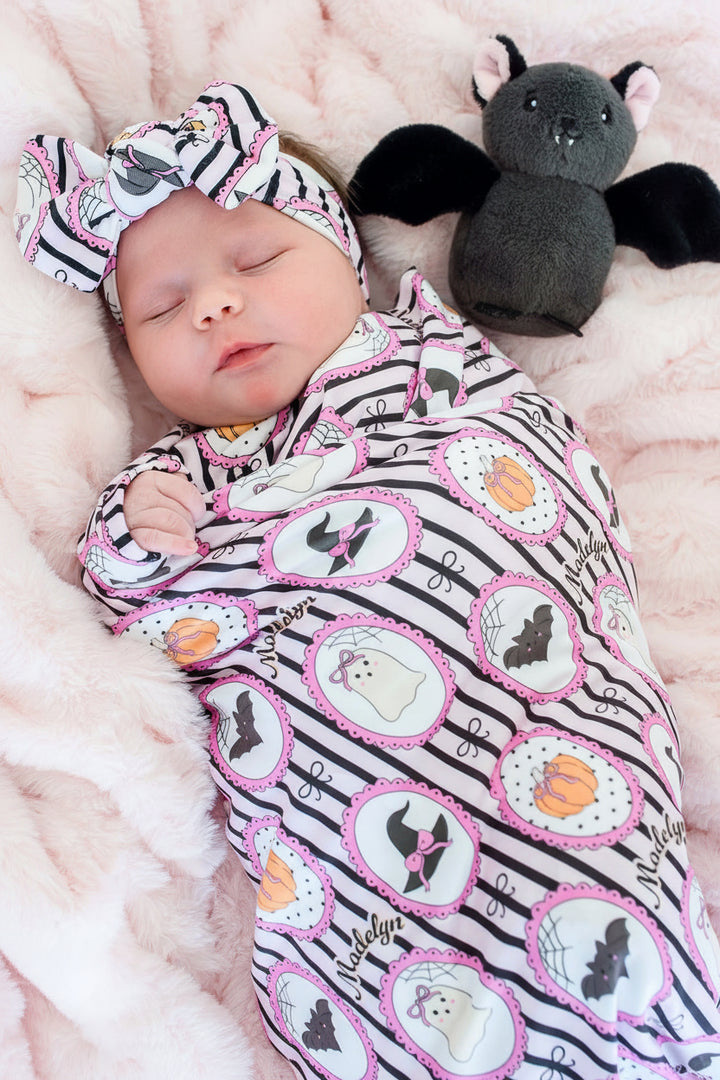 A baby swaddled in a black and white striped blanket with pink-edged circular Halloween motifs, wearing a matching headband, lying on a pink blanket next to a plush bat toy.