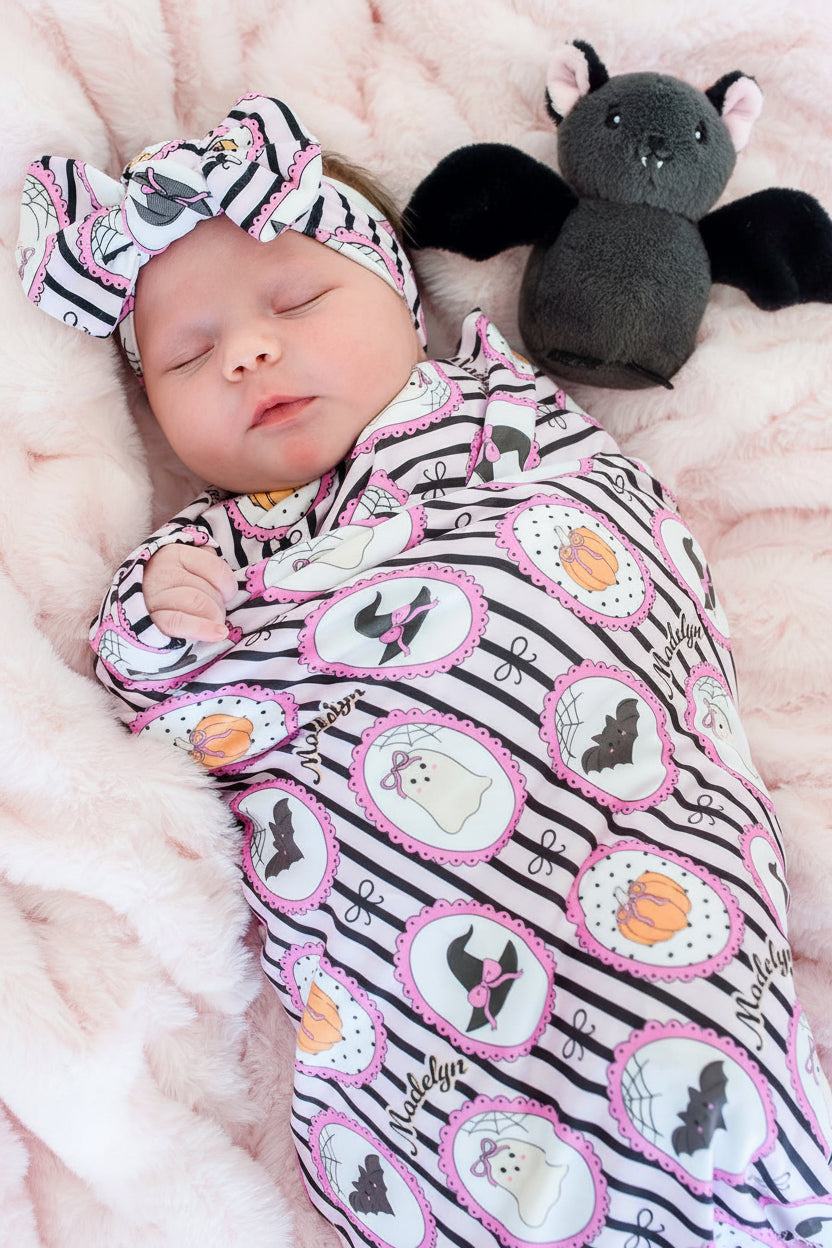 A baby swaddled in a black and white striped blanket with pink-edged circular Halloween motifs, wearing a matching headband, lying on a pink blanket next to a plush bat toy.