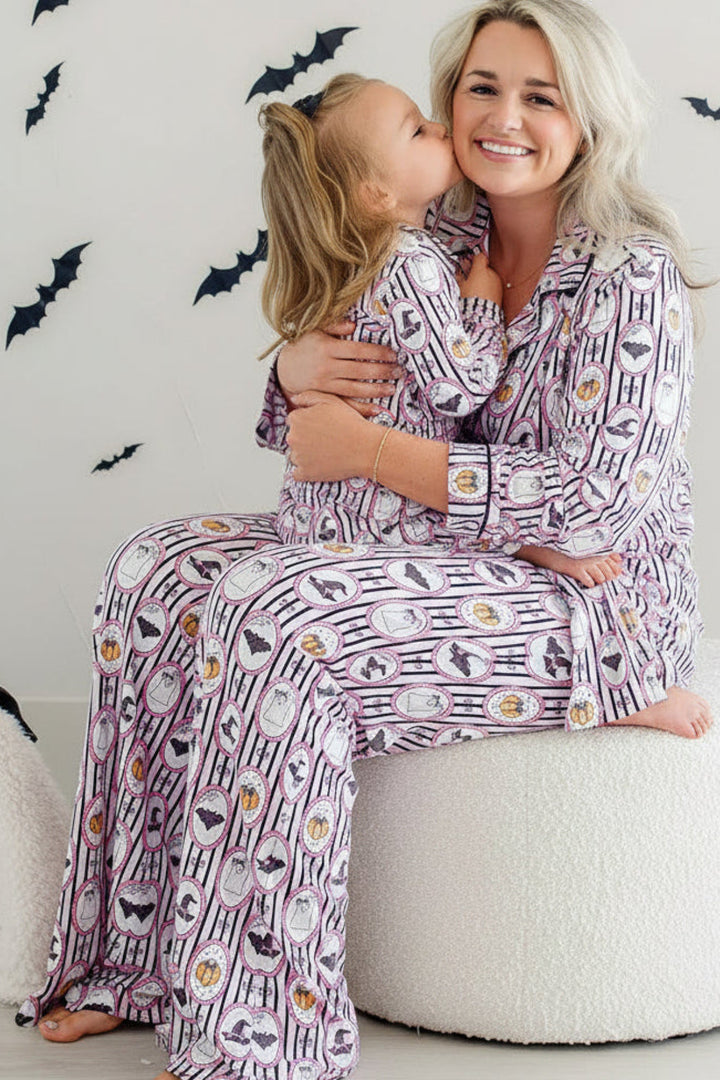 A woman and a young girl wearing matching purple and white patterned pajamas sit together on a round white pouf, with black bat decals on a light wall behind them.