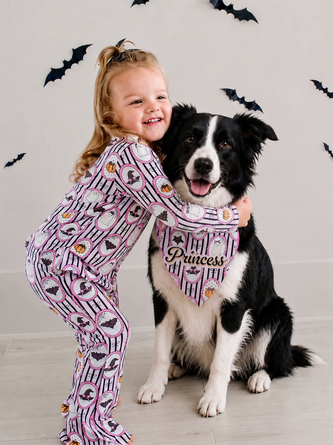 A young girl in Halloween-print pajamas hugging a black and white dog wearing a pink 'Princess' bandana, with bat decorations on the wall behind them.