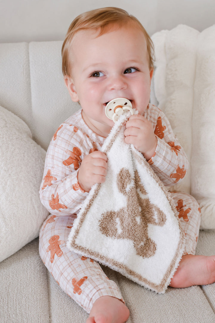 Smiling baby in Caden Lane gingerbread man pajamas holding a tan and white gingerbread man lovie security blanket with pacifier