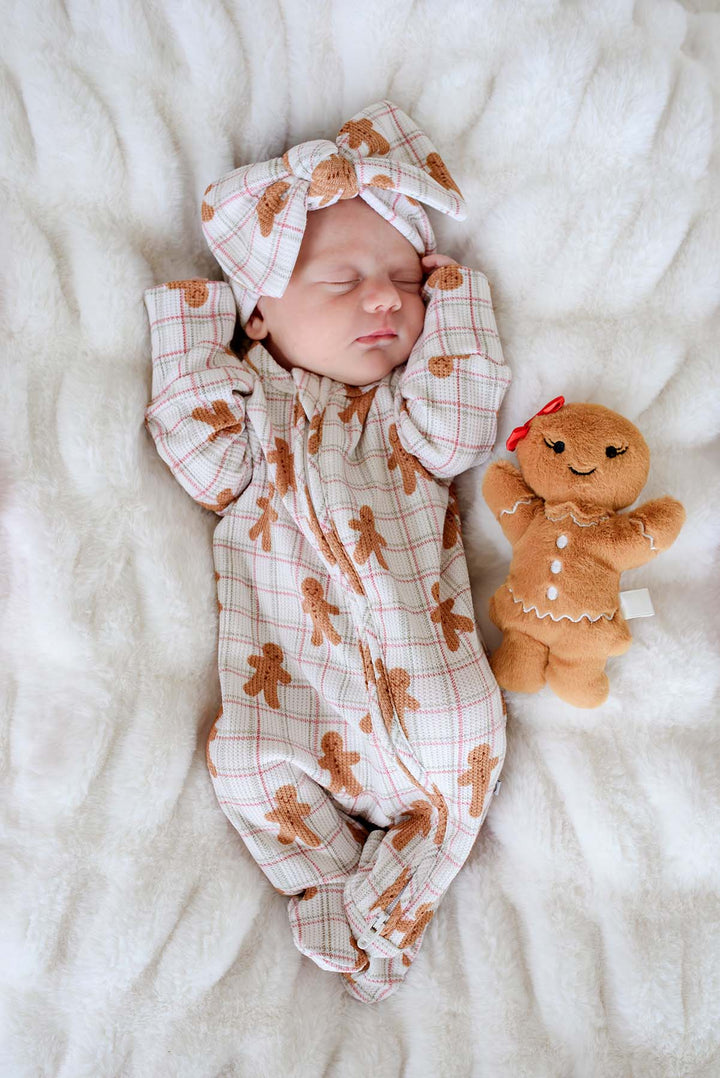 Caden Lane newborn wearing a bamboo convertible zipper romper and matching bow headwrap in a festive gingerbread cookie print, resting beside a gingerbread plush on a cozy blanket—perfect for baby's first Christmas, holiday photos, and winter gifting.