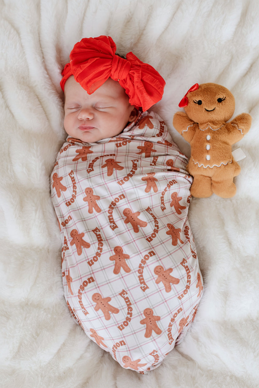 sleeping newborn swaddled in a personalized Caden Lane gingerbread print christmas swaddle blanket with a red bow headband