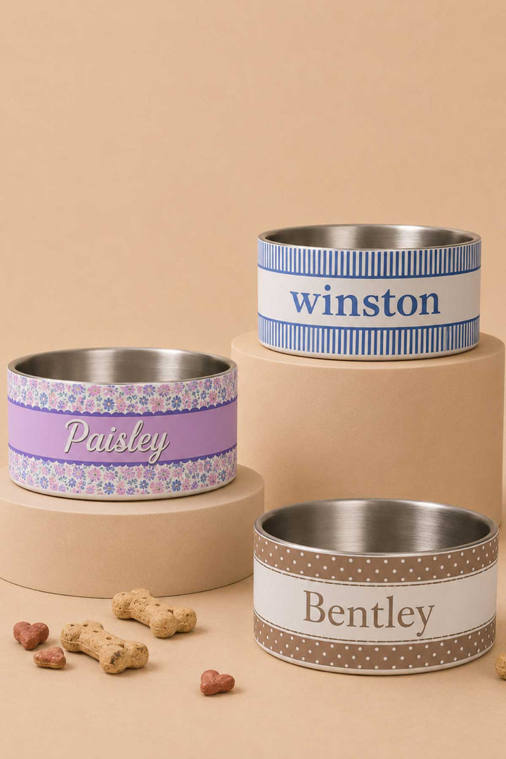 Three personalized pet bowls displayed on risers with dog treats: "Paisley" in the Ribbon Garden floral print, "Winston" in a blue stripe, and "Bentley" in a brown polka dot pattern