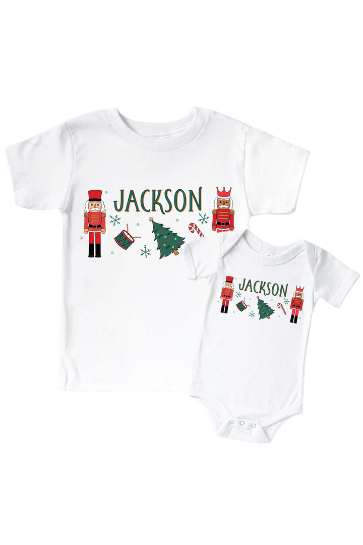 Caden Lane's matching t-shirt and onesie personalized with the name "Jackson," both featuring red toy soldier nutcrackers, a Christmas tree, drum, and candy cane, laid flat together for a sibling set.