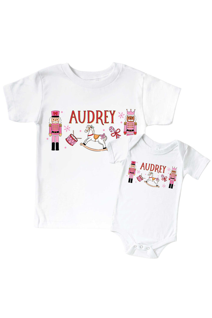 Caden Lane's matching t-shirt and onesie personalized with the name "Audrey," both featuring pink toy soldier nutcrackers, a rocking horse, drum, and candy cane, laid flat together for a sibling set.