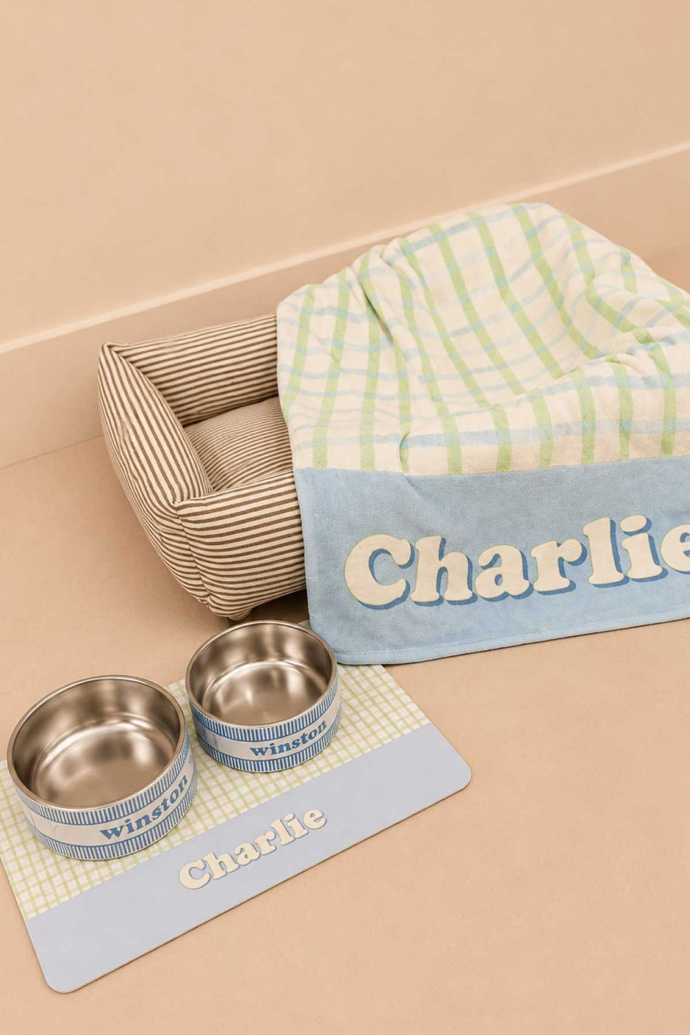 Lifestyle photo showing a picnic plaid pet blanket draped over a wooden pet bed frame with a personalized placemat labeled "Charlie" and matching double bowls labeled "Winston" in front