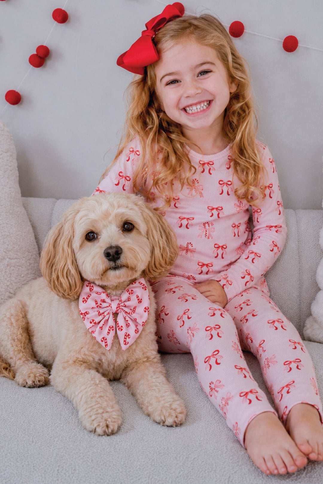 Girl in pink holiday pajamas smiling next to a goldendoodle wearing a Lane and Leash candy cane print bandana.