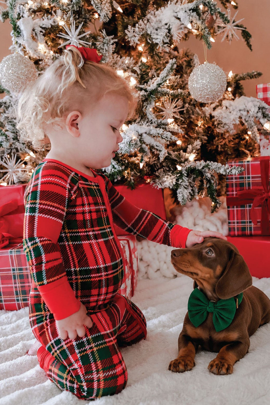 Caden Lane toddler wearing a red and green plaid bamboo Christmas romper while petting a dachshund puppy in a green bow tie, perfect for matching family Christmas pajamas and holiday memories.